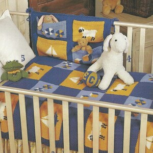 Puede incluir: Un juego de cuna de bebé tejido en azul y amarillo con un parachoques, una manta y una falda a juego. La manta y el parachoques presentan cuadrados con diseños de veleros, ovejas y geométricos. Un perro de peluche blanco y una rana verde están en la cuna.