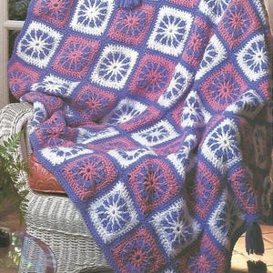 May include: A handmade crochet blanket draped over a wicker chair. The blanket features a repeating pattern of squares in white, pink, and purple, with a floral design in each square. The blanket has a purple border and tassels.