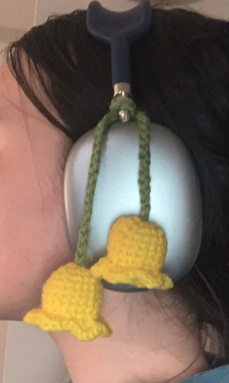 May include: A pair of yellow crocheted bells hanging from a blue and green cord attached to a silver object. The bells are attached to a silver object with a blue handle.