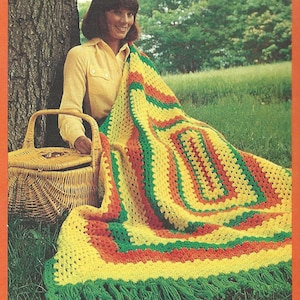 May include: A woman sits on the grass with a picnic basket and a crocheted blanket in yellow, green, and orange stripes. The blanket has fringe on the bottom.