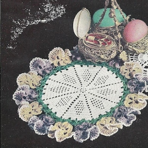 Pode incluir: Um tapete de crochê branco com uma borda verde e um design floral. O tapete é rodeado por flores de crochê em tons de roxo, amarelo e verde.