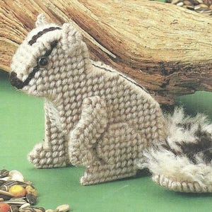 May include: A handcrafted chipmunk figurine made from beige plastic canvas with dark brown stitched stripes along the head and back. The figure features a small black nose, dark eyes, and a fluffy, textured tail with brown and white faux fur.