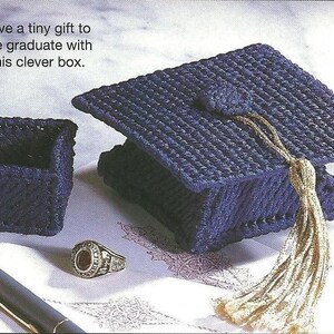 May include: A dark blue plastic canvas graduation cap box with a gold tassel, a small square dark blue container, and a silver ring. The text reads: Give a tiny gift to the graduate with this clever box. The items are arranged on a white surface.