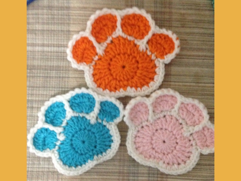 May include: Three crocheted paw-shaped coasters in orange, blue, and pink. Each coaster has a white border. The coasters are handmade and suitable for pet lovers. The coasters are approximately 4 inches in diameter.
