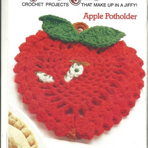 Pode incluir: Um porta-copos de maçã vermelho em crochê com folhas verdes. O porta-copos é feito com um padrão de ponto simples e tem um caule branco em crochê.