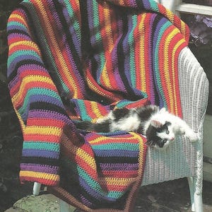 Può includere: Una coperta all'uncinetto con strisce verticali in colori vivaci tra cui rosso, giallo, viola, verde acqua e nero. La coperta è drappeggiata su una sedia in vimini bianco e un piccolo gattino bianco e nero è sdraiato sulla superficie.