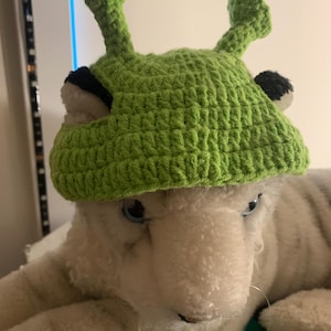 May include: A bright green crocheted hat featuring two upright antenna-like protrusions on top and two small black circular accents on the sides, designed to resemble a creature with stalks, worn on a white plush toy with blue eyes.