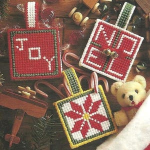 Könnte beinhalten: Drei quadratische Kunststoff-Stickerei-Ornamente mit weihnachtlichen Motiven. Ein rotes Ornament zeigt den weißen Schriftzug JOY, ein weißes Ornament den roten Schriftzug NOEL mit goldenen Glöckchen und ein weiteres weißes Ornament einen rot-gelben Weihnachtsstern.