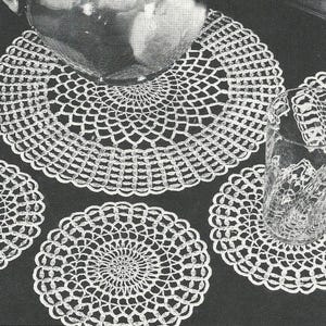 May include: White crocheted doilies and a large placemat are arranged on a dark surface. The doilies are circular with intricate openwork patterns. A glass pitcher and drinking glasses are also visible, suggesting a dining or decorative setting.