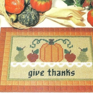 May include: A rectangular needlepoint mat featuring a central orange pumpkin flanked by two red apples and two green pears, all resting on a white scalloped lace trim. The text "give thanks" is stitched in black below the fruit design.