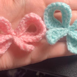 May include: Two handmade crochet bows, one light pink and one light blue, are displayed. The bows are small, with a classic bow shape, and appear to be made of soft yarn. The bows are shown against a skin-toned background.