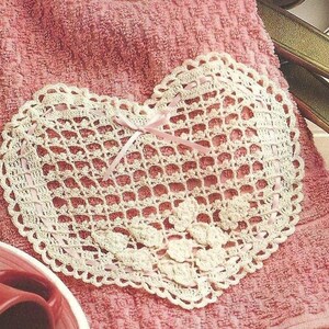 May include: A cream-colored, heart-shaped doily with a delicate, open-weave design. The doily features a scalloped edge and is adorned with small floral accents and a pink ribbon. It rests on a pink towel.