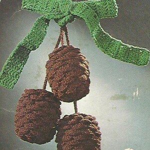 May include: A handmade crochet ornament featuring three brown, textured, cone-shaped elements suspended from a green bow. The bow is a vibrant green color, and the cones have a detailed, textured appearance. The overall theme is a rustic, handcrafted decoration.