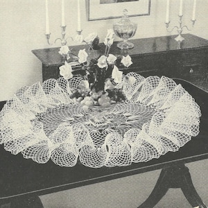 May include: A white, oval-shaped, crocheted doily with a ruffled edge, displayed on a dark table. The doily is decorated with a floral arrangement and fruit. A dark wooden cabinet and candlesticks are in the background.