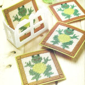 Könnte beinhalten: Ein Set aus quadratischen Untersetzern aus Kunststoff-Stramin mit einem grünen und gelben Froschmotiv und braunem Rand, aufbewahrt in einem weißen Halter. Jeder Untersetzer zeigt ein gesticktes Froschdesign auf weißem Grund.