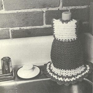 May include: A crocheted bottle cover in the shape of a dress, with a white bodice and a dark-colored skirt. The skirt has a white and dark-colored scalloped trim. The bottle cover is sitting on a sink.