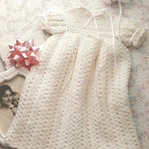 May include: A cream-colored, crocheted christening gown set with a matching bonnet and booties. The gown features a detailed, openwork design and delicate ribbon accents. The image also includes a framed photograph and a pink bow.