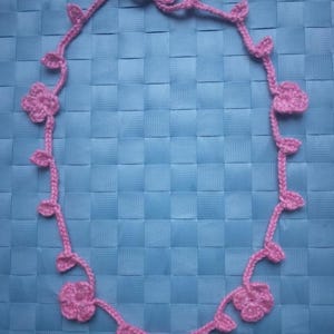 Puede incluir: Un collar rosa hecho a mano con ganchillo, con un diseño floral y de hojas. El collar está hecho con una cadena delicada y adornado con múltiples flores y hojas de ganchillo. El collar se muestra sobre una superficie azul texturizada.