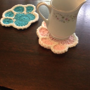 May include: Two handmade crochet paw print coasters, one teal and white, the other pink and white, sit on a dark wooden table. A white ceramic creamer with floral detailing rests on the pink coaster. The coasters add a whimsical touch.