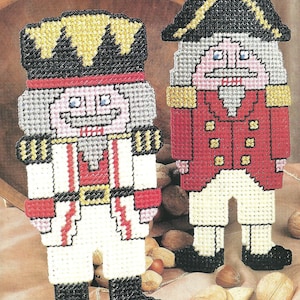 May include: Two plastic canvas nutcracker figures featuring cross-stitch patterns. One figure wears a white uniform with red accents and a black hat, while the other wears a red uniform with white pants and a black hat with gold trim.