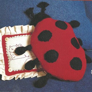 May include: A knitted ladybird toy with black spots and a matching black head and legs rests on a small pillow with a cat design. The pillow has a red border and ruffled edges. The background is a blue denim fabric.