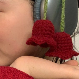 May include: A person wearing a red knitted sweater with a black design and a pair of silver headphones with a green crocheted flower attached.