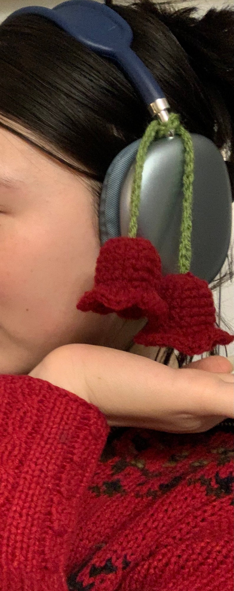 May include: A person wearing a red knitted sweater with a black design and a pair of silver headphones with a green crocheted flower attached.