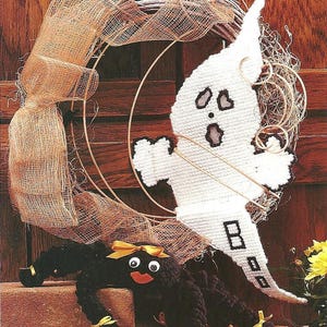 Pode incluir: Uma guirlanda decorativa com um fantasma de tela plástica branca com bordas pretas e a palavra BOO bordada em preto. O fantasma está preso a uma guirlanda de videira adornada com fita de malha bege e uma aranha de pelúcia preta.