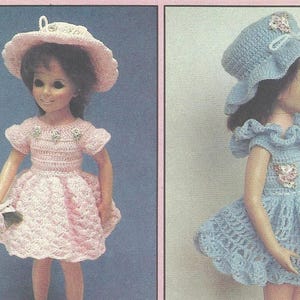 May include: Two dolls wearing handmade crochet dresses and hats. One doll is in a light pink dress and hat, holding a bouquet. The other doll is in a blue dress and hat. Both outfits feature floral accents.