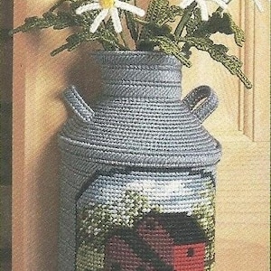 May include: A gray, crocheted vase with a rural scene featuring a red barn and white fence. The vase holds crocheted yellow and white flowers with green leaves. The vase has two handles.