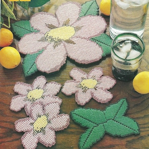 Può includere: Un set di sottobicchieri a punto croce a forma di fiori rosa con centri gialli e foglie verdi. La collezione comprende un fiore grande, due fiori più piccoli e un sottobicchiere a forma di foglia, tutti con un visibile motivo a griglia.