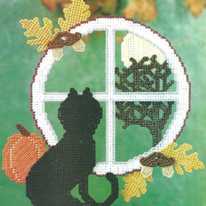 Può includere: Una decorazione in tela plastica raffigurante un gatto nero seduto davanti a una cornice circolare bianca. La scena include una zucca arancione, foglie di quercia gialle, ghiande marroni e la sagoma di un albero verde scuro.