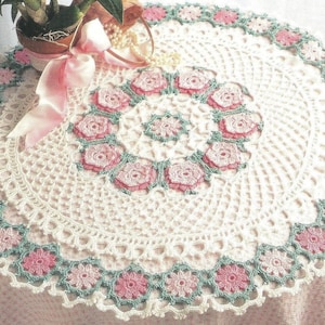 Könnte beinhalten: Runder, weiß gehäkelter Tischläufer mit floralem Design. Der Tischläufer hat einen zentralen Ring aus rosa Rosen und einen Rand aus rosa und grünen Blumen. Das filigrane, spitzenartige Muster verleiht ihm eine zarte Note.
