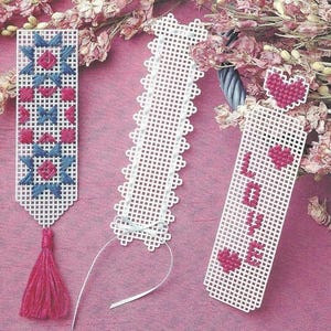 Könnte beinhalten: Drei rechteckige Lesezeichen aus Kunststoff-Stramin. Das linke zeigt geometrische Sternmuster in Blau und Pink mit einer pinkfarbenen Quaste. Das mittlere ist schlicht weiß mit gewelltem Rand und einem dünnen weißen Band. Das rechte zeigt das Wort LOVE in Pink.