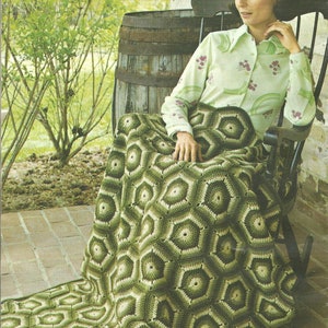 May include: A woman sits in a rocking chair on a porch, wrapped in a green and white crocheted blanket with a hexagonal pattern. The blanket is made of multiple shades of green and white yarn.