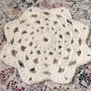 May include: A white crocheted doily with a starburst design. The doily is made with a delicate, lacy stitch and has a scalloped edge.