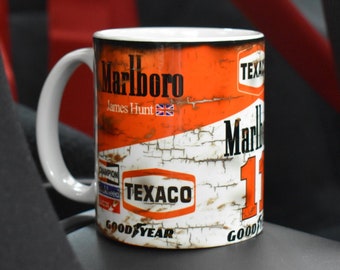 Puede incluir: Una taza de cerámica blanca con un diseño desgastado que presenta el logotipo de Marlboro, el nombre de James Hunt y los logotipos de Texaco y Goodyear. La taza tiene un asa blanca y está sobre un fondo oscuro.