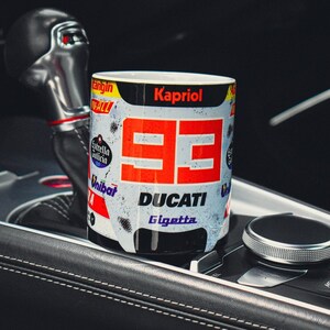 May include: A white ceramic mug with a red and black racing design. The mug features the number "99" in red, the word "DUCATI" in black, and the word "Gigetta" in black. The mug also has the word "Kapriol" in black at the top.