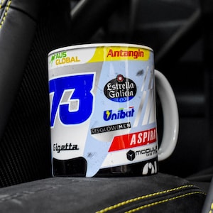 May include: A white ceramic mug with a blue number 73, the words "Gigetta", "Antangin", "MSGLOWMEN", "ASPIRA", and "Estrella Galicia 001", and a red and white logo. The mug is sitting in a black leather car seat.