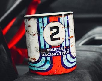 Puede incluir: Una taza cilíndrica de metal envejecido con un diseño de carreras. La taza presenta el número "2" en un círculo blanco, junto con las palabras "MARTINI RACING-TEAM" y rayas azules, rojas y blancas. Se encuentra en un interior oscuro.