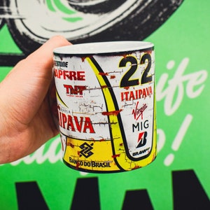 May include: A white ceramic mug with a vintage racing car design. The mug features a yellow and red color scheme with the number 22, the words "Itaipava" and "Banco do Brasil", and the logos of "TNT Energy Drink", "Virgin", "MIG", and "Brawn GP".
