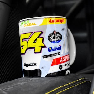 May include: White ceramic mug with a racing car design. The mug features the number 54 in yellow, the words "Gigetta" and "Aspira", and various sponsors' logos, including "Antangin", "Estrella Galicia", and "Unibat".