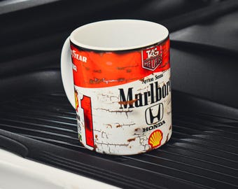 Puede incluir: Una taza de cerámica blanca con un diseño rojo y blanco con el logotipo de Marlboro, el logotipo de Honda y el número 1. La taza tiene un asa blanca y está sobre una superficie negra.