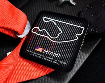 May include: A black and white track map of the Miami International Autodrome race track with the text "MIAMI" and "MIAMI INTERNATIONAL AUTODROME, FLORIDA 19 CORNERS - 5.412 KM (3.363 MI)" printed on it. The map is on a black carbon fiber background.