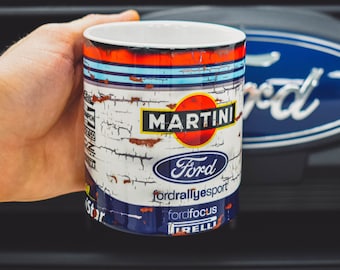Puede incluir: Una taza de cerámica blanca con un diseño azul, rojo y blanco. La taza presenta el logotipo de Martini Racing, el logotipo de Ford y el texto "Fordrallyesport" y "Fordfocus".