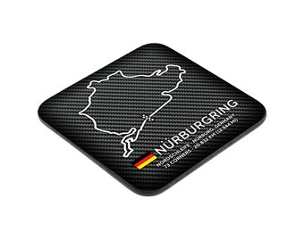 May include: Black square coaster with a white outline of the Nürburgring race track. The coaster has a carbon fiber pattern and the text "Nürburgring, Nordschleife - Nürburg, Germany, 73 Corners - 20.832 km (12.944 mi)".