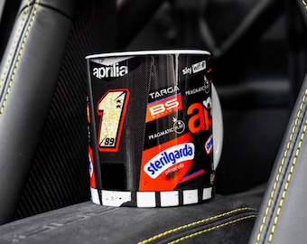 May include: A black and white ceramic mug with a variety of colourful stickers and logos, including "Aprilia", "Targa BS", "Pragmatic Play", "Sterilgarda", and "Sky Wifi".