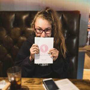 May include: A young person wearing glasses is holding a pink and white card with the text "sd&wn" and a pink feather logo. The card is in front of their face. The person is sitting at a table with a glass of liquid and a cell phone.