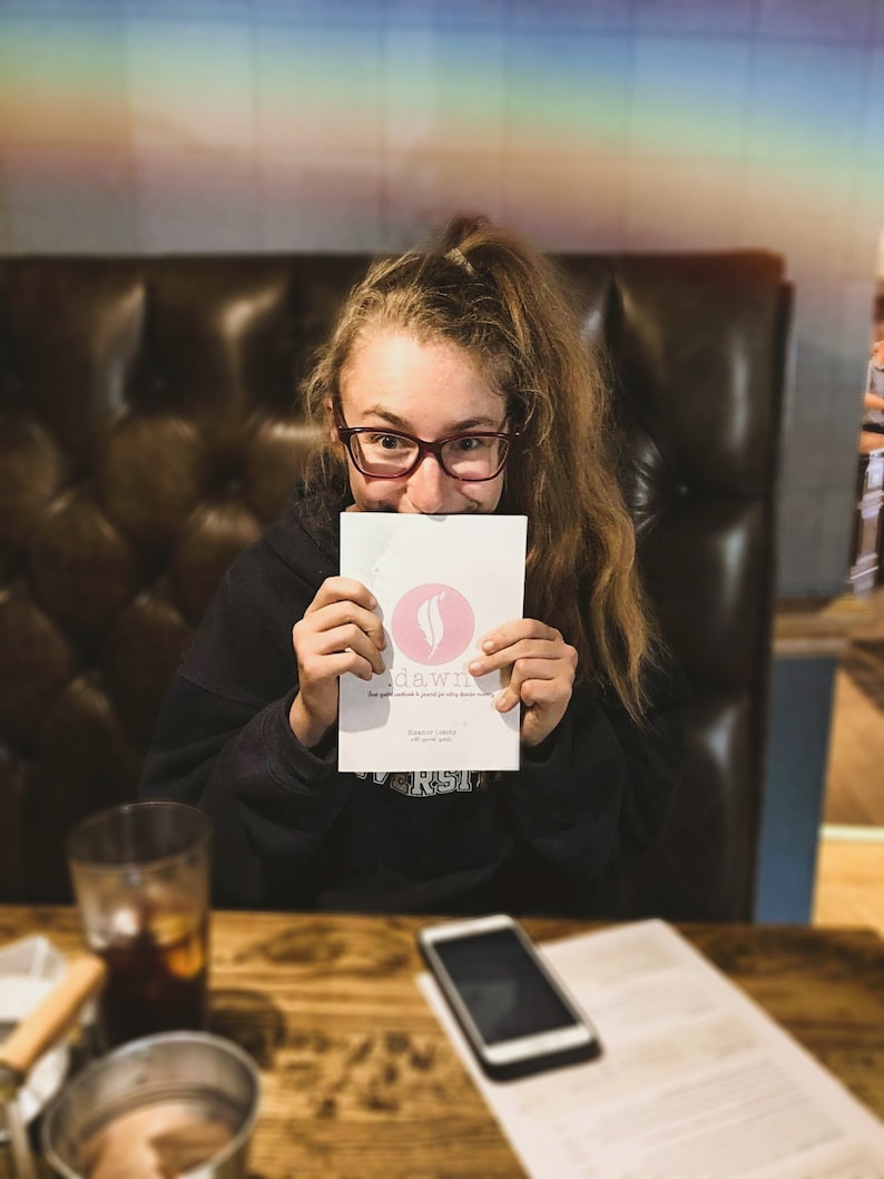 May include: A young person wearing glasses is holding a pink and white card with the text "sd&wn" and a pink feather logo. The card is in front of their face. The person is sitting at a table with a glass of liquid and a cell phone.