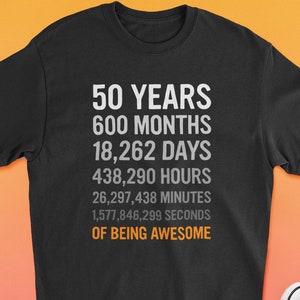 May include: Black t-shirt with white text that reads "50 Years 600 Months 18,262 Days 438,290 Hours 26,297,438 Minutes 1,577,846,299 Seconds of Being Awesome". The t-shirt is laid out on a pink and orange gradient background with white shorts and pink sneakers.